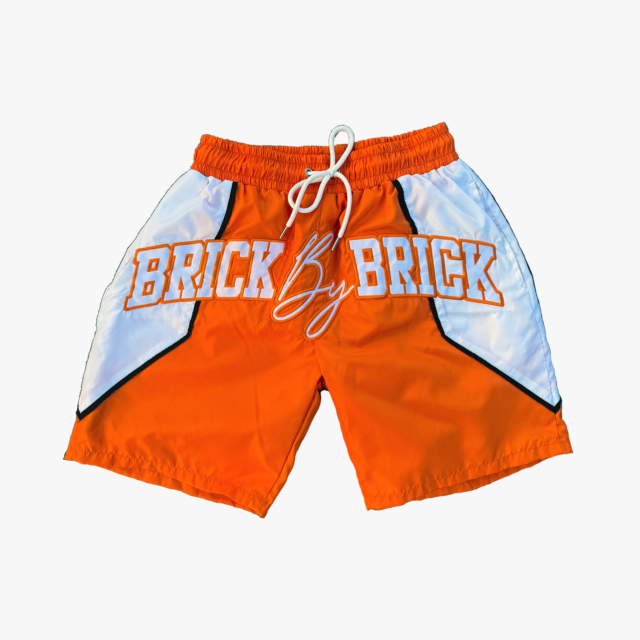 Orange organic nylon shorts with 'BRICK BY BRICK' text on a white background